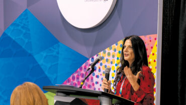 A woman with long dark hair speaks at a podium during the Advanced Textiles Expo, colorful backdrop with geometric patterns visible.