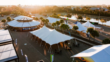Spacious outdoor venue featuring white tents and a circular building, bathed in warm sunlight, near a waterway.