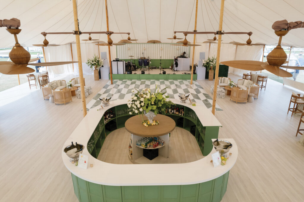 Elegant tent interior featuring a green and white checkerboard floor, a central bar adorned with flowers, and stylish seating arrangements.