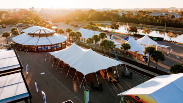 Aerial view of a tent expo with white tents and a circular building, set in a sunny outdoor venue with palm trees and a nearby water body.