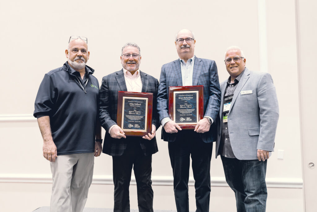 Four men stand together, holding framed awards, in front of a neutral backdrop. They are dressed in business attire, smiling.