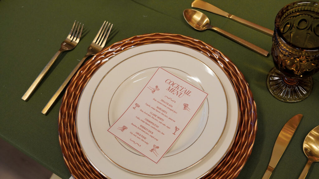 Elegant table setting with a green tablecloth, gold utensils, a decorative plate, and a cocktail menu in the center. A brown glass sits beside.