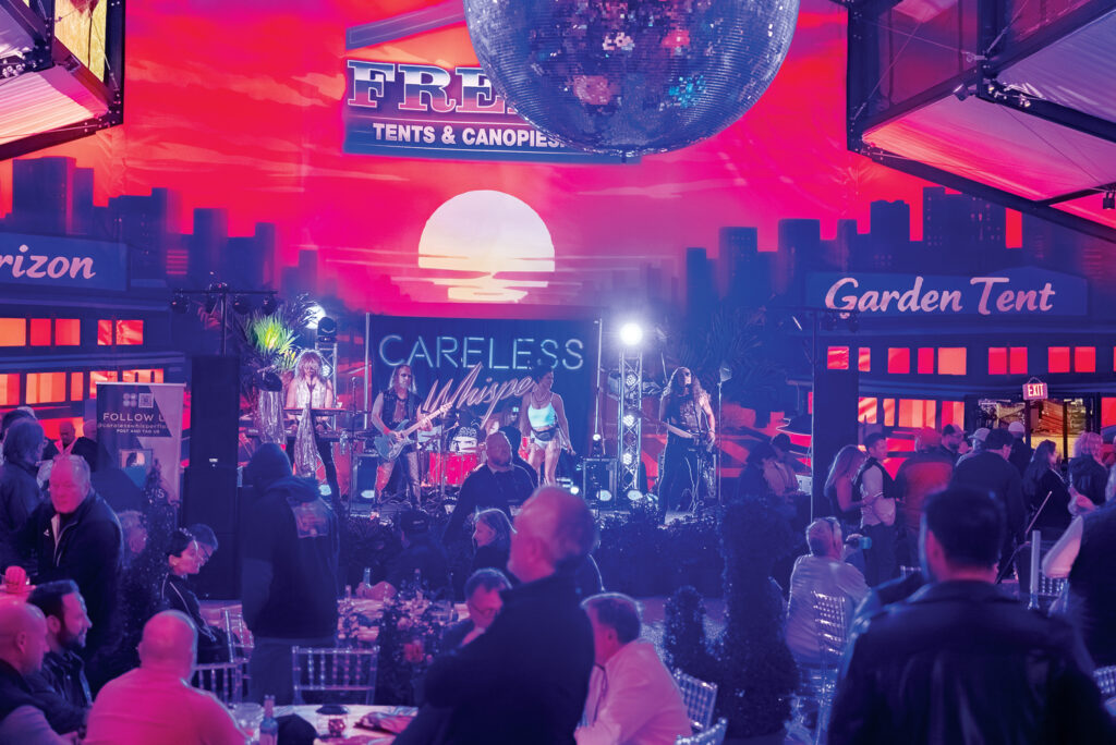 A lively band performs on stage under a disco ball, vibrant sunset backdrop, with attendees seated at tables enjoying the event.