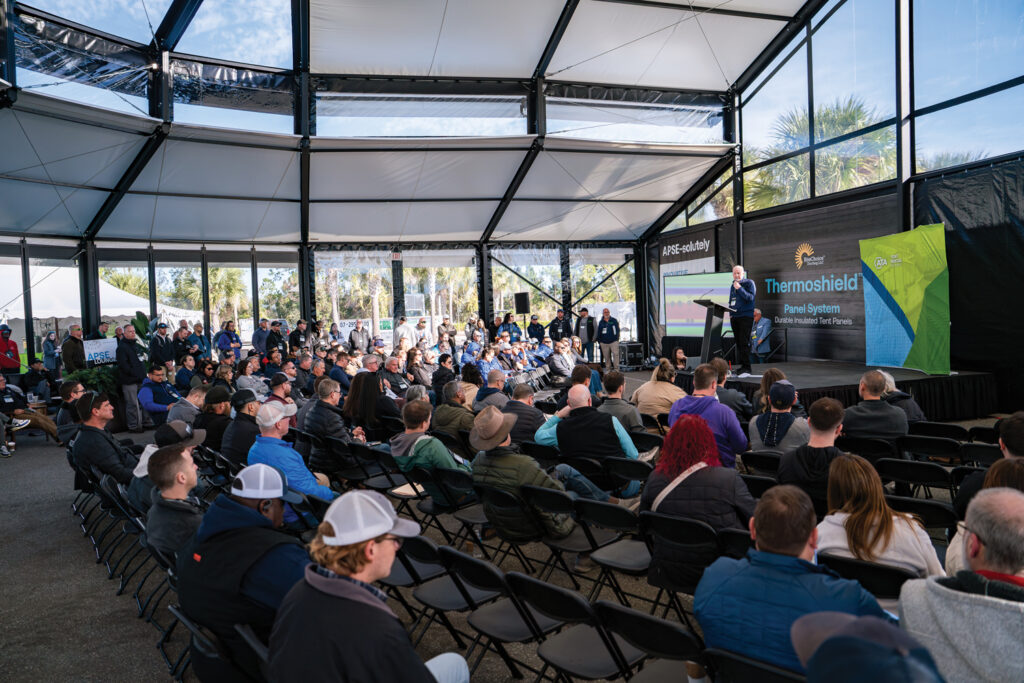 A crowd attentively listens to a speaker in a large tent; rows of chairs face a stage with banners highlighting the Thermoshield system.