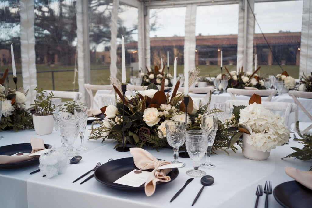 Elegant table setting in a tent, featuring dark plates, crystal glassware, white floral centerpieces, and white candles amidst greenery.
