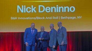 Four men in formal attire stand on stage under a bright yellow backdrop, celebrating Nick Deninno with an award.