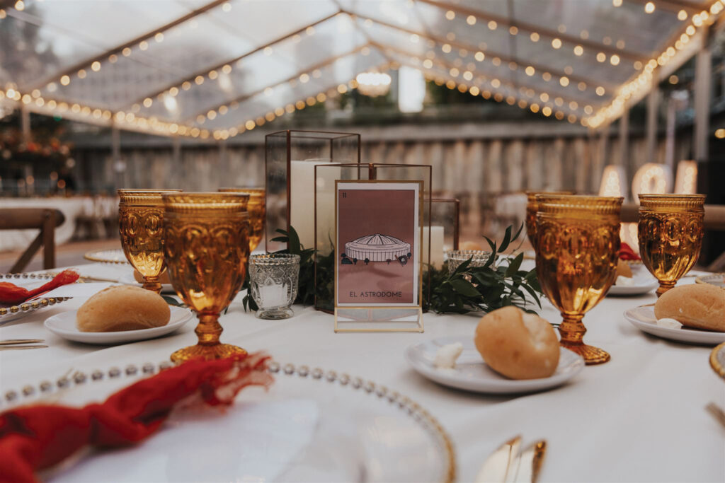 Elegant table setting with golden goblets, a centerpiece featuring a menu card, and rolls on white plates, all under soft string lights.