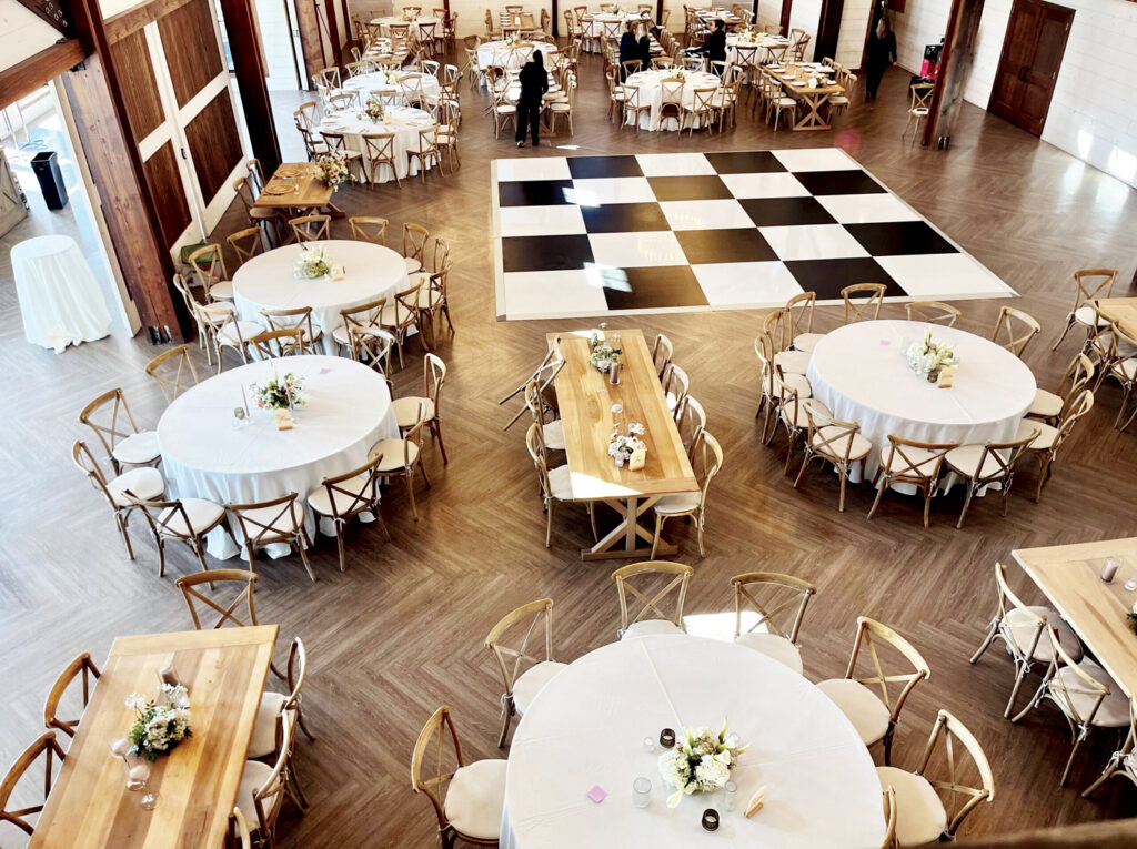 Bright venue featuring circular tables dressed in white linens, wooden accents, and a large black-and-white dance floor in the center.
