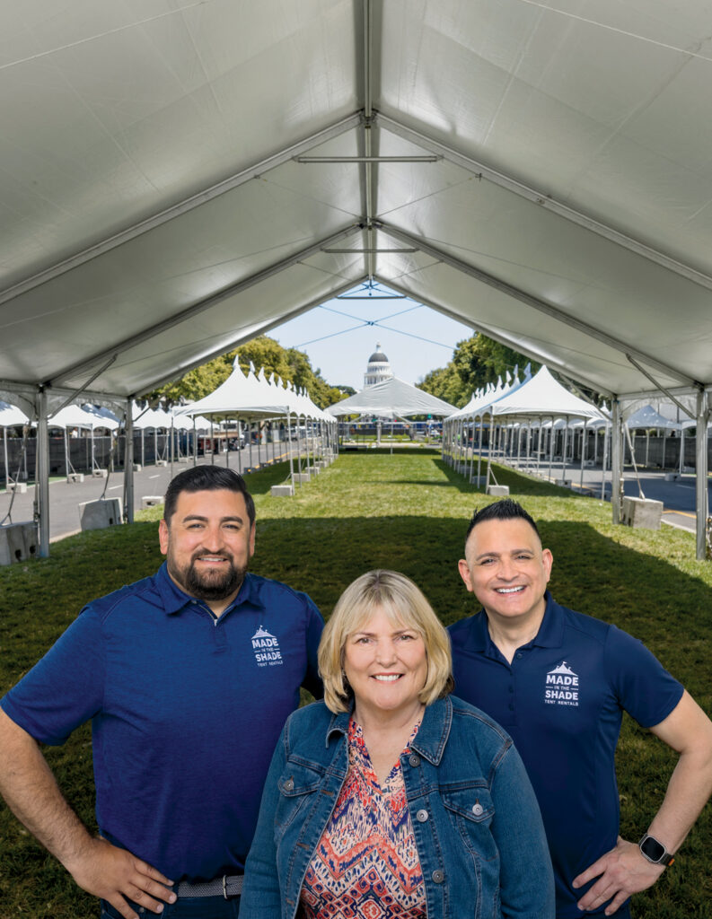 Three smiling individuals stand in front of a row of white tents under a clear blue sky, with a green lawn and a building in the background.