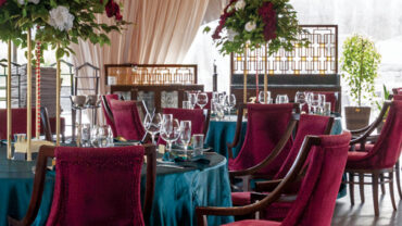 Elegant dining setup featuring emerald green tables, deep red chairs, and lush floral centerpieces, surrounded by large windows.