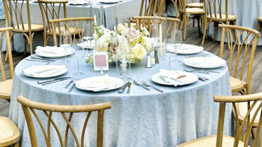 Elegant dining setup with light blue tablecloths, wooden chairs, and floral centerpieces on round tables, creating a sophisticated atmosphere.