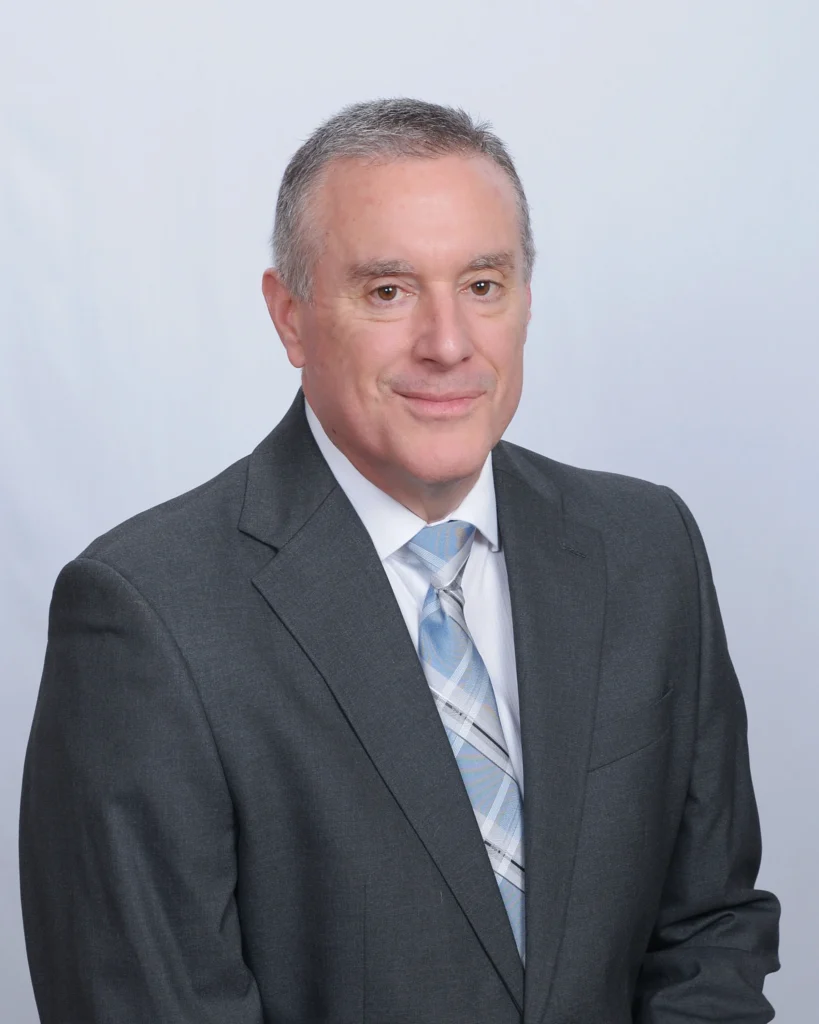 Professional headshot of a man, Lyle Shipley, in a dark grey suit with a light blue and white plaid tie, set against a plain white background.