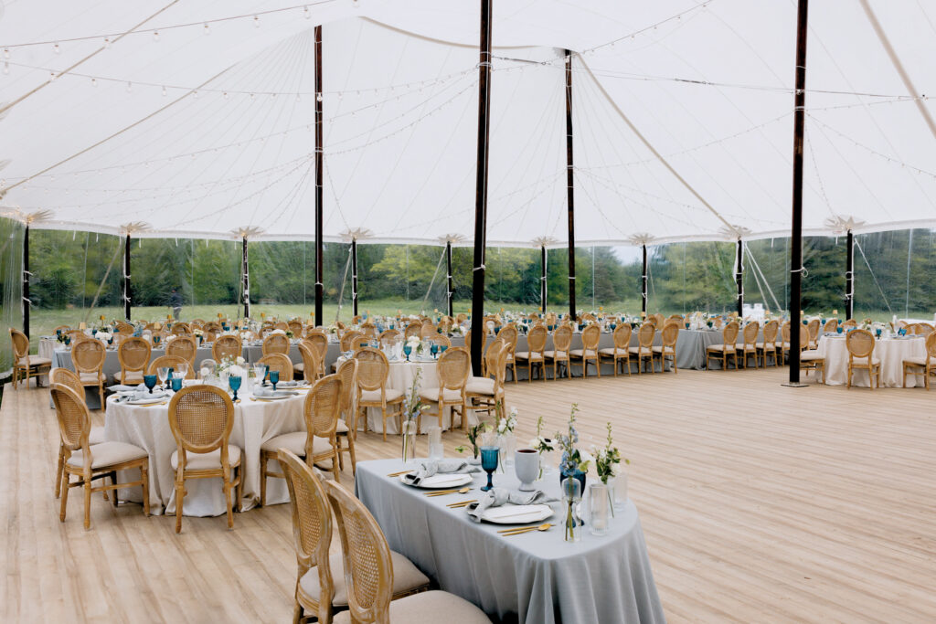 Elegant tented event space featuring tables with light blue tablecloths, wicker chairs, glassware, and floral centerpieces, under a white canopy.