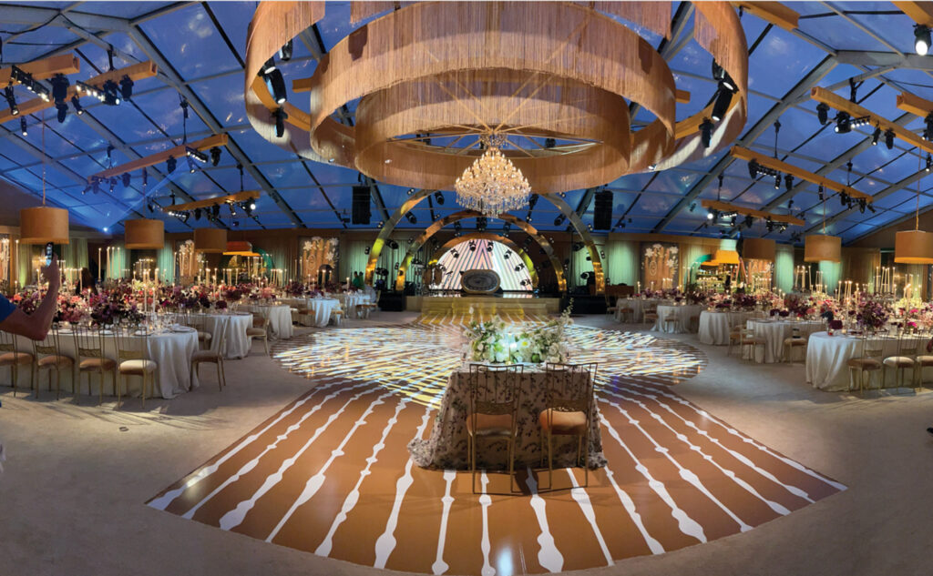 Elegant event space featuring round tables adorned with white tablecloths and floral centerpieces, under a decorated, illuminated canopy.