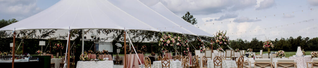 A large white tent decorated with floral arrangements, surrounded by tables set for an event in an outdoor setting under cloudy skies.
