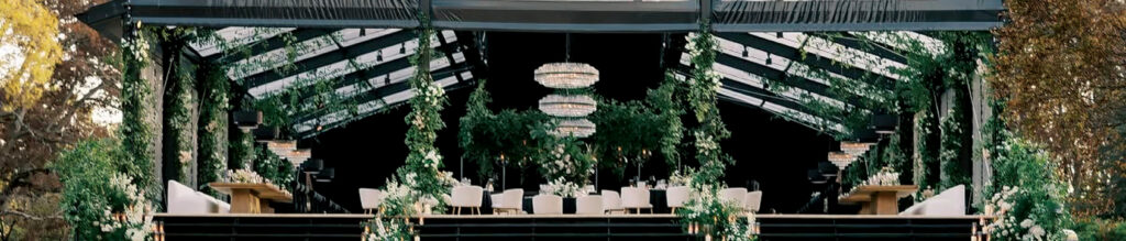 A decorated indoor space featuring greenery, two chandeliers, and elegant seating arranged for a gathering or event.