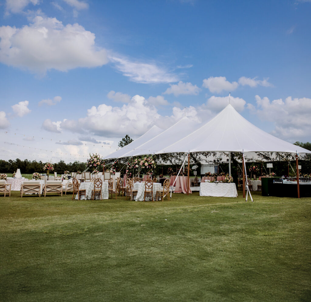 A large white tent with elegant table arrangements under a blue sky, surrounded by lush green grass and floral decorations.