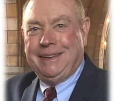 Smiling man in a dark suit with a red tie, set against a softly blurred historic interior background.