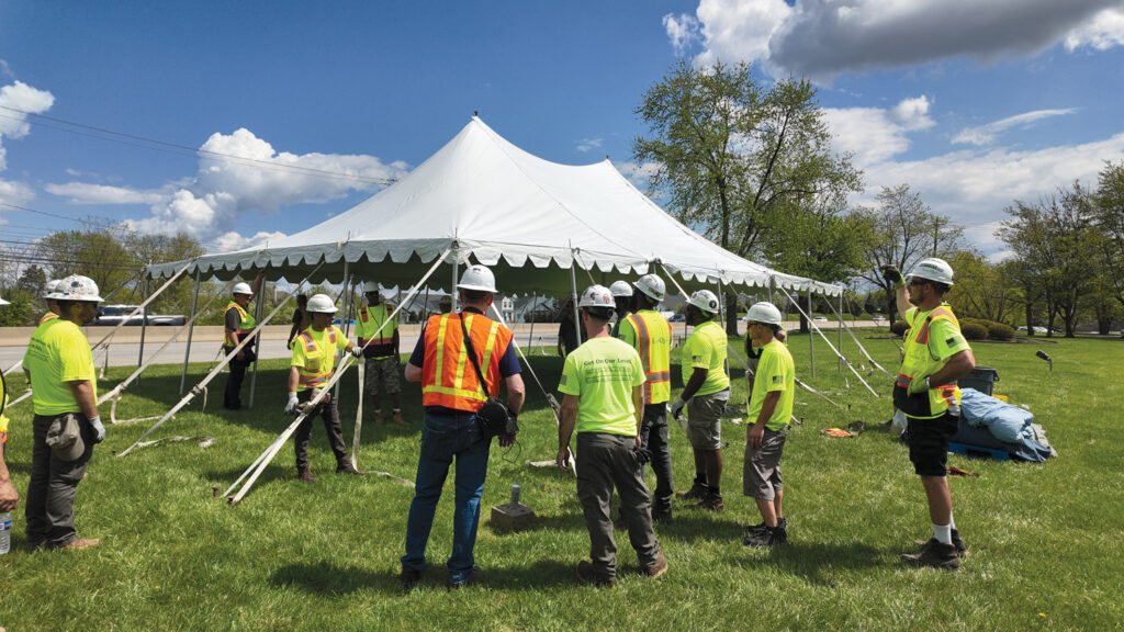 A group of individuals in safety vests and helmets gathers around a large white tent on a grassy area under a clear blue sky.