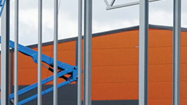 Construction scene featuring a blue crane lifting materials against a backdrop of an orange warehouse and steel framework.