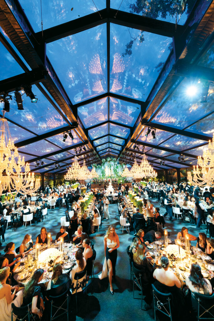 A grand indoor event with a transparent roof, chandeliers, and elegantly dressed guests seated at long tables under a starlit sky.