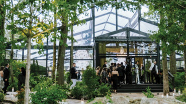 A glass-walled venue surrounded by greenery, with guests mingling outside, warmly lit by chandeliers inside.