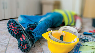A construction worker lies on the ground in a yellow safety vest, with a yellow hard hat and tools scattered around, suggesting an accident scene.