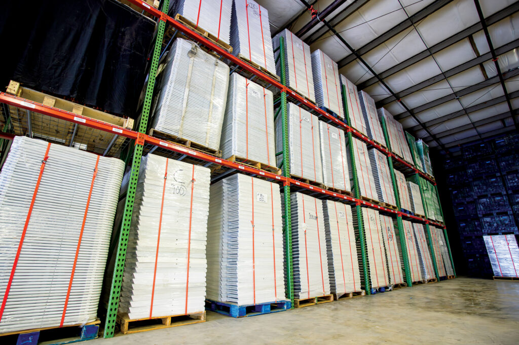Stacks of white panels on pallets are organized in a warehouse, with green shelving and industrial lighting above.