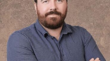 Man with a beard wearing a blue checked shirt, crossing his arms, standing against a textured brown background.