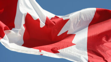 Canadian flag fluttering against a clear blue sky, prominently displaying the red and white colors with a central maple leaf emblem.