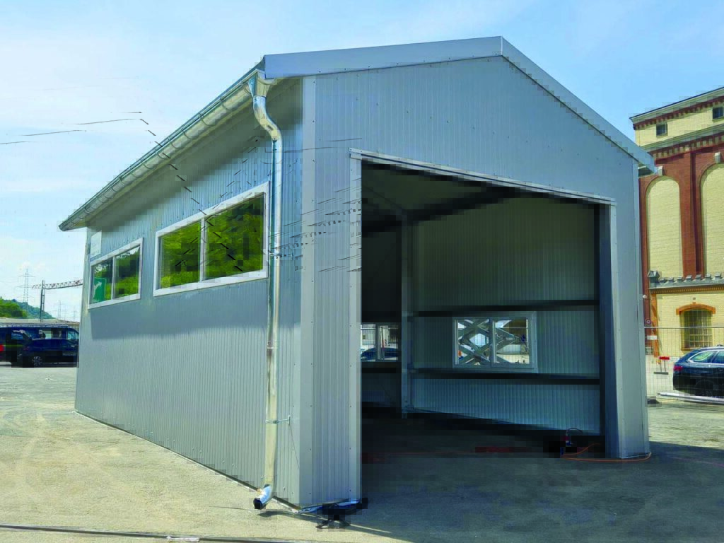 A large, gray metal building with a gabled roof and wide open door, set against a clear blue sky and a paved area.