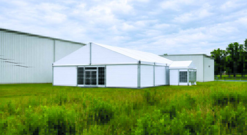 Large white tent structure on green grass, with a backdrop of grey buildings under a cloudy sky.
