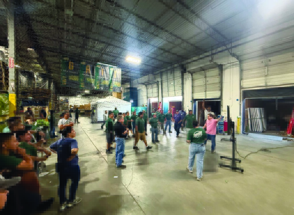 A group of people in green shirts gathers in a spacious warehouse, engaged in an activity, with colorful banners hanging above.