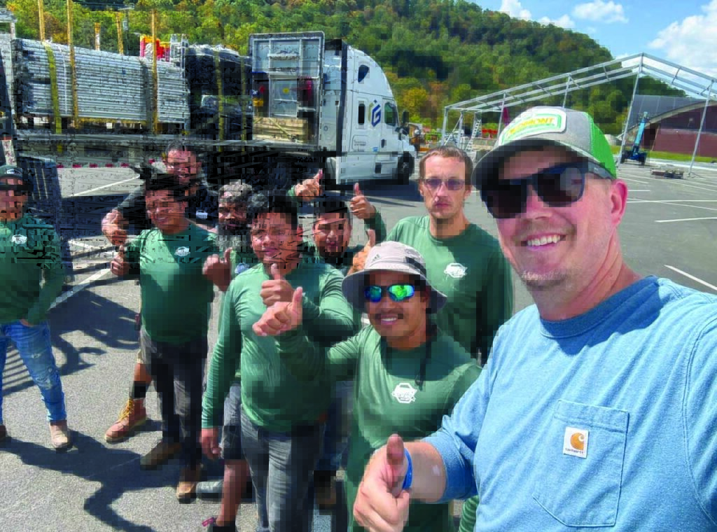A group of smiling workers in green shirts gives thumbs up, with a truck and construction materials in the background and trees on a hill.