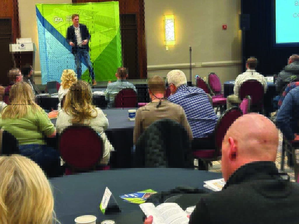 A speaker presents on stage to an audience seated at round tables, with a green and blue backdrop featuring a tent design.
