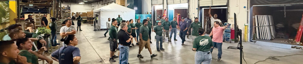 A diverse group of people in green shirts gathers in a spacious warehouse, engaged in discussion near equipment and storage areas.