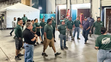 A diverse group of people in green shirts gathers in a spacious warehouse, engaged in discussion near equipment and storage areas.