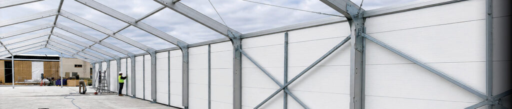 A large, partially constructed tent with a steel frame and white fabric walls, showing workers and an open sky in the background.