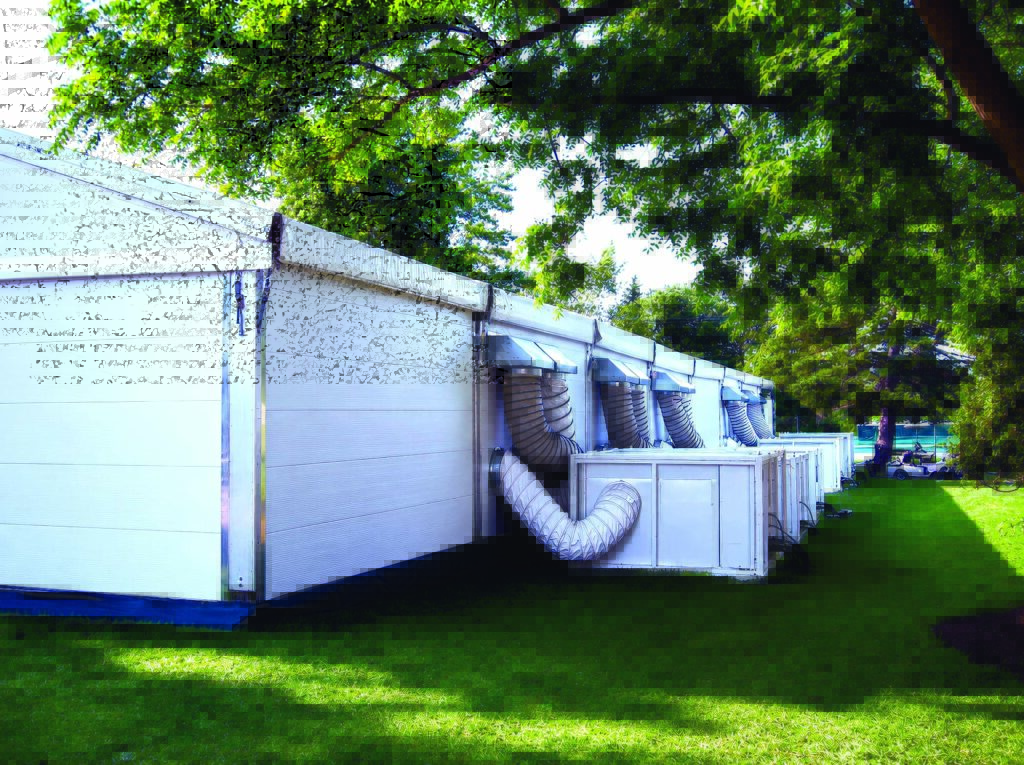 White modular structures with ventilation ducts extend from one side, set on green grass under dappled sunlight from surrounding trees.