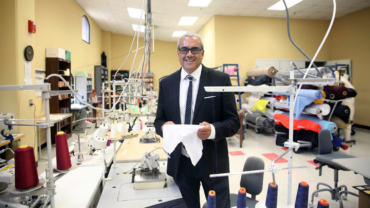 A smiling man in a suit stands in a textile workshop, holding a piece of fabric, surrounded by sewing machines and colorful rolls of material.